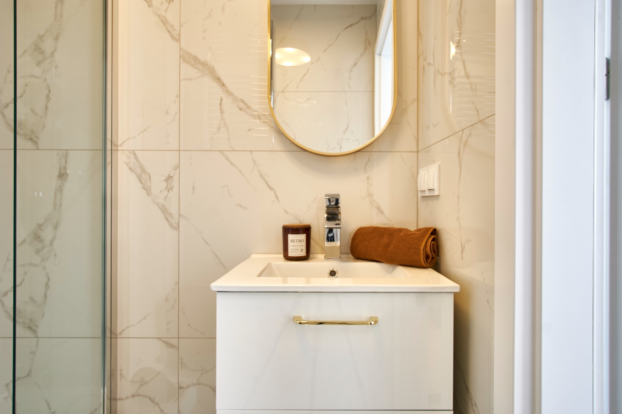Elegancka łazienka z umywalką, lustrem w złotej ramie i marmurowymi płytkami. Świeca zapachowa i ręcznik dodają przytulności. Minimalistyczny design.