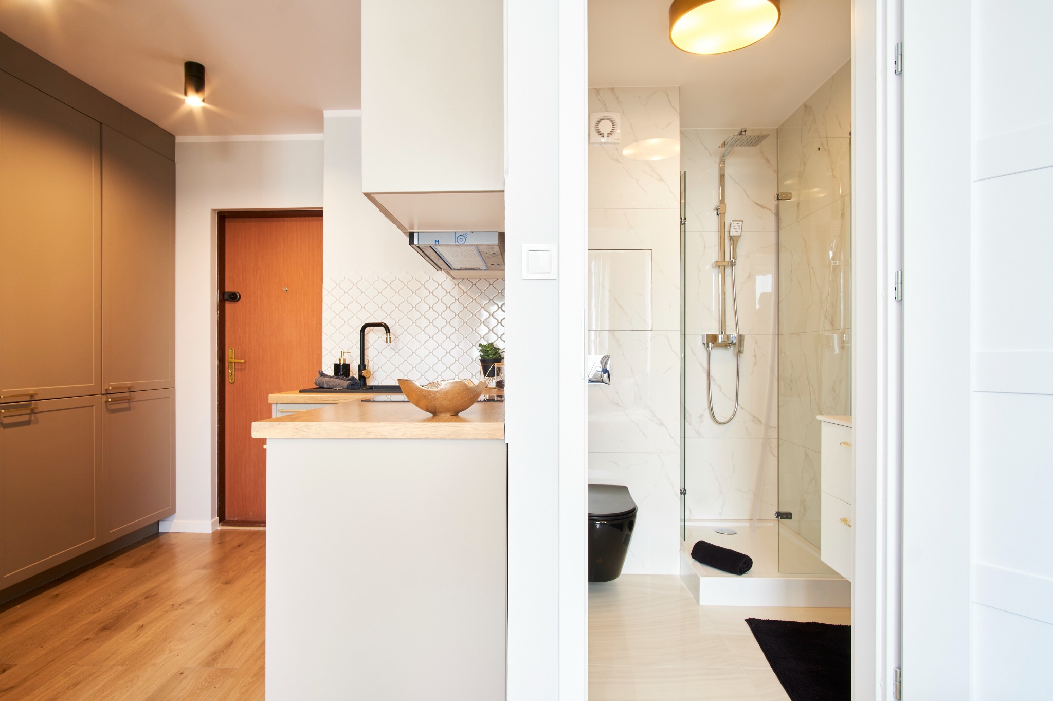 Nowoczesne wnętrze: fragment kuchni z drewnianym blatem i widok na łazienkę z kabiną prysznicową, czarną toaletą i marmurowymi płytkami. Oświetlenie sufitowe.
