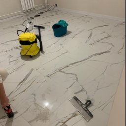 CLEAN INTERIOR 24 TOMASZ KASZUBOWSKI - Błyszcząca, marmurowa podłoga po czyszczeniu, z widocznym żółtym odkurzaczem parowym, niebieskim wiadrem i narzędziem do usuwania zabrudzeń. Widoczny fragment osoby.