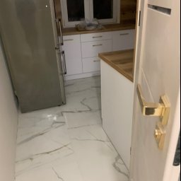 CLEAN INTERIOR 24 TOMASZ KASZUBOWSKI - Nowoczesna kuchnia z marmurową podłogą i białymi szafkami, widziana z perspektywy otwartych drzwi. Lodówka ze stali nierdzewnej po lewej stronie.