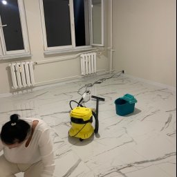 CLEAN INTERIOR 24 TOMASZ KASZUBOWSKI - Kobieta w trakcie czyszczenia marmurowej podłogi za pomocą profesjonalnego odkurzacza i wiadra w jasnym, pustym pokoju z oknami i grzejnikami.