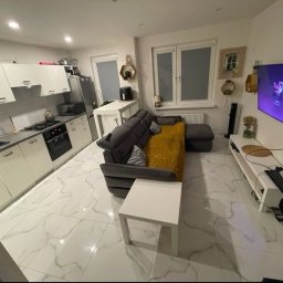 CLEAN INTERIOR 24 TOMASZ KASZUBOWSKI - Jasne wnętrze mieszkania z aneksem kuchennym, szarą sofą z żółtą poduszką i białymi meblami. Marmurowe płytki podłogowe. Nowoczesny design z elementami dekoracyjnymi na ścianach.