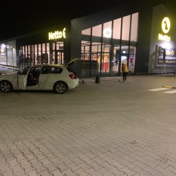 CLEAN INTERIOR 24 TOMASZ KASZUBOWSKI - Nocne sprzątanie kostki brukowej przed sklepem Netto. Osoba w żółtej kurtce zamiata, obok stoi biały samochód z otwartymi drzwiami i bagażnikiem.