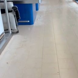 CLEAN INTERIOR 24 TOMASZ KASZUBOWSKI - Wnętrze komercyjne z rzędem niebieskich stanowisk roboczych z metalowymi blatami, widok na czystą, jasną podłogę i odległą postać w tle. Perspektywa z dołu.