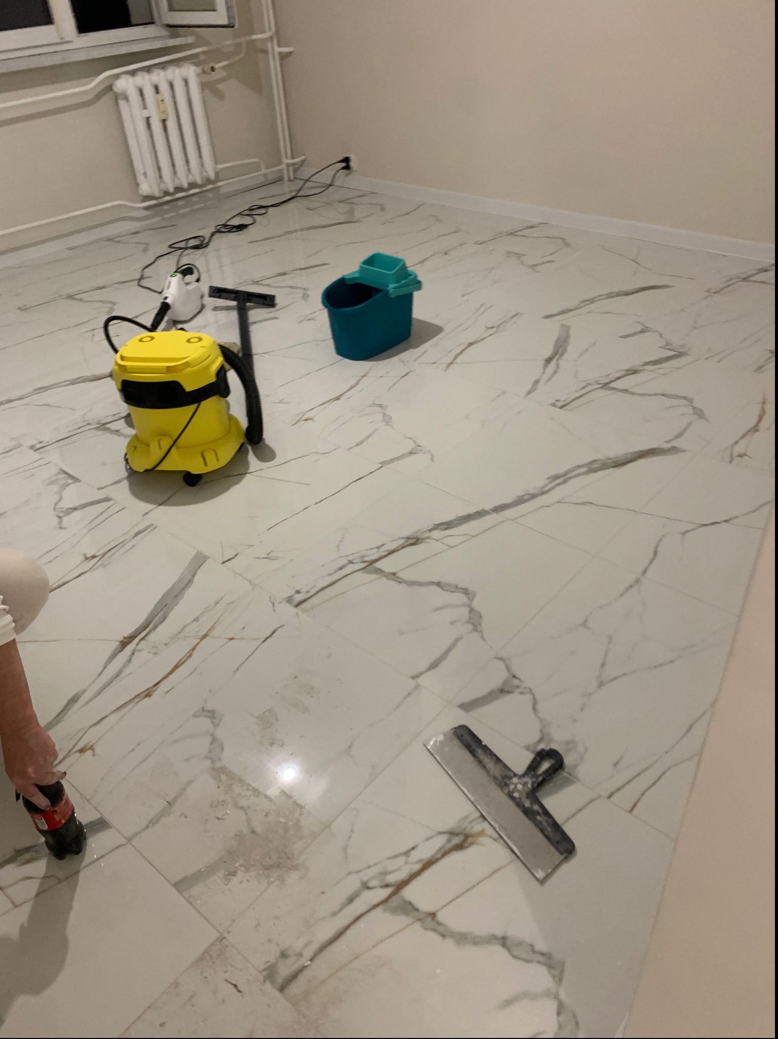 Błyszcząca, marmurowa podłoga po czyszczeniu, z widocznym żółtym odkurzaczem parowym, niebieskim wiadrem i narzędziem do usuwania zabrudzeń. Widoczny fragment osoby.