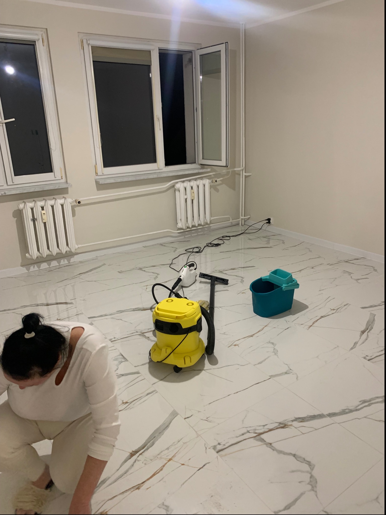 Kobieta w trakcie czyszczenia marmurowej podłogi za pomocą profesjonalnego odkurzacza i wiadra w jasnym, pustym pokoju z oknami i grzejnikami.