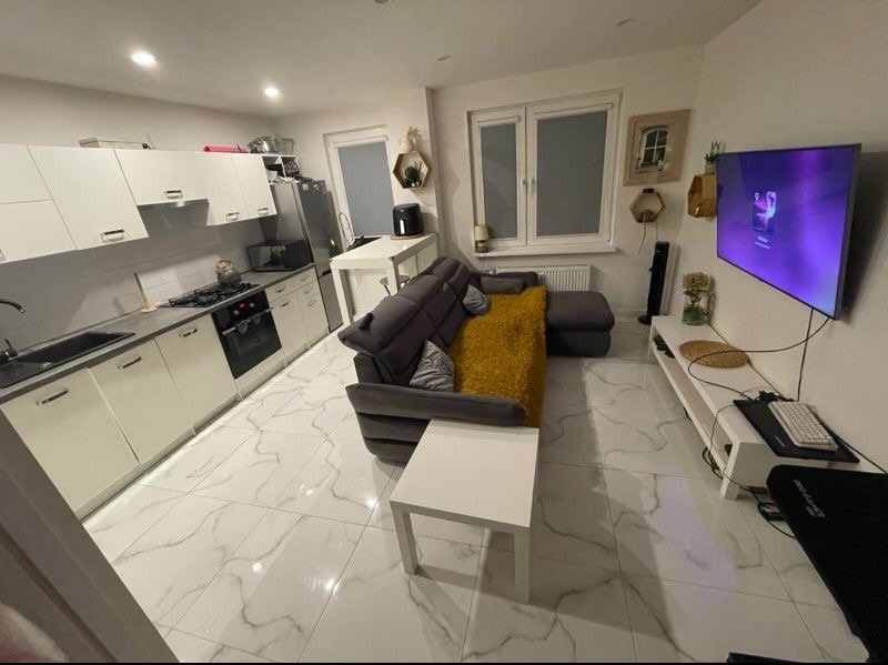 Jasne wnętrze mieszkania z aneksem kuchennym, szarą sofą z żółtą poduszką i białymi meblami. Marmurowe płytki podłogowe. Nowoczesny design z elementami dekoracyjnymi na ścianach.