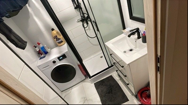 Wnętrze łazienki z pralką, kabiną prysznicową i umywalką. Na pralce stoją środki czystości. Widok z perspektywy drzwi, z lekkim obrotem kadru.