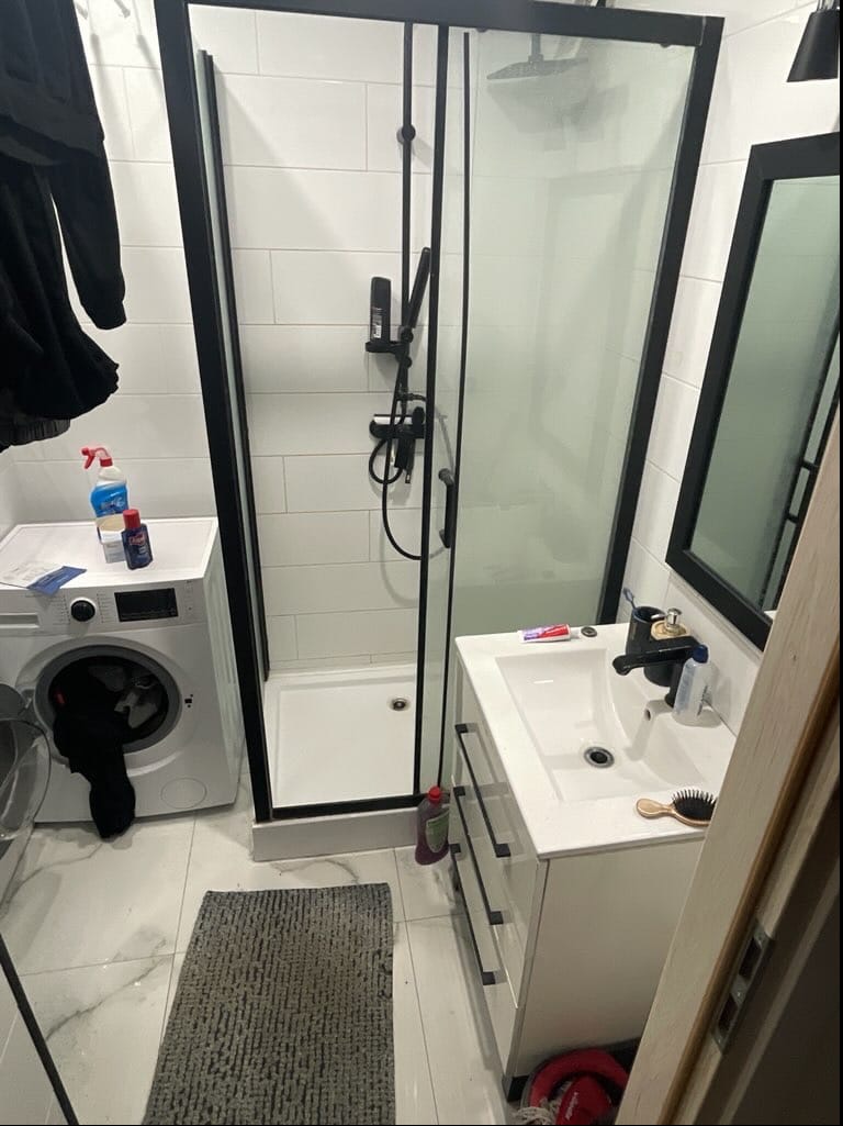 Nowoczesna łazienka z kabiną prysznicową w czarnej ramie, białą umywalką z szafką i pralką. Minimalistyczny design, białe płytki i szary dywanik.