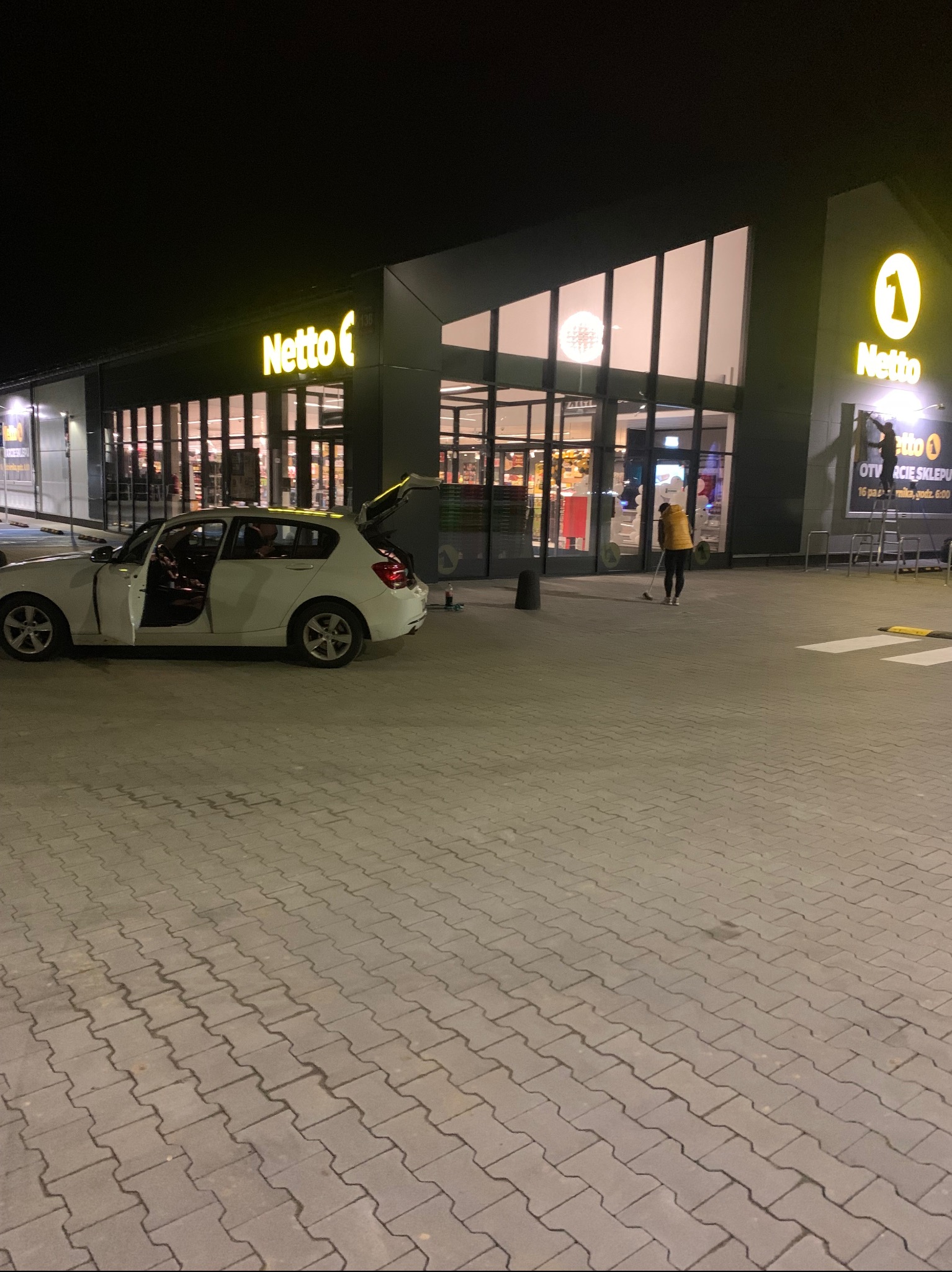 Nocne sprzątanie kostki brukowej przed sklepem Netto. Osoba w żółtej kurtce zamiata, obok stoi biały samochód z otwartymi drzwiami i bagażnikiem.