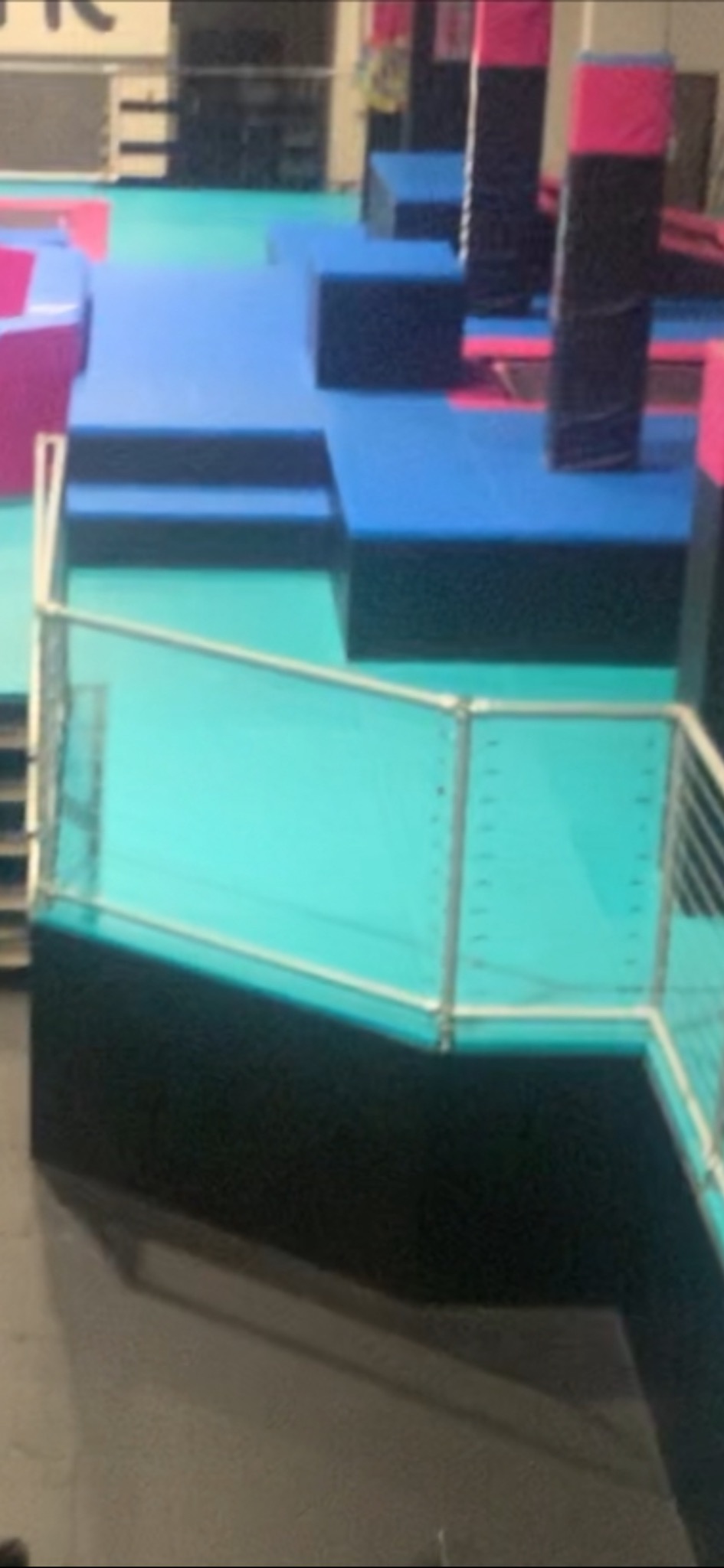 Kolorowa konstrukcja z platform i barierki w sali zabaw. Elementy w odcieniach niebieskiego, różowego i czarnego. Widok z góry na fragment placu zabaw.