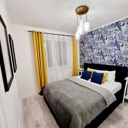 Patrico Build and Design Patryk Sysiak - Nowoczesna sypialnia z tapetą w tropikalne liście, żółtymi zasłonami i szarą narzutą na łóżku. Widoczne drewniane panele podłogowe i lampa wisząca.