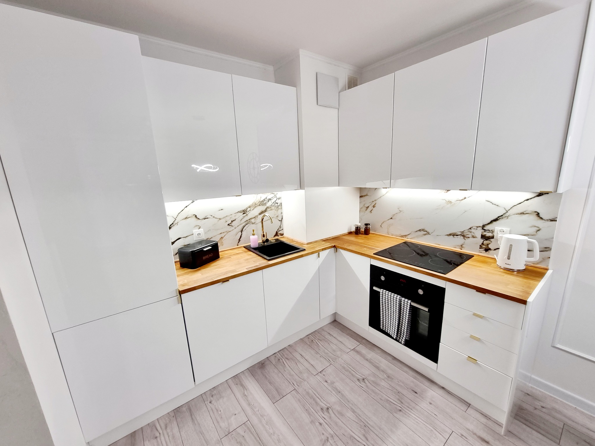 Biała, nowoczesna kuchnia z drewnianym blatem i marmurowym backsplashiem. Widok na zabudowę kuchenną z piekarnikiem i płytą indukcyjną. Minimalistyczny design.