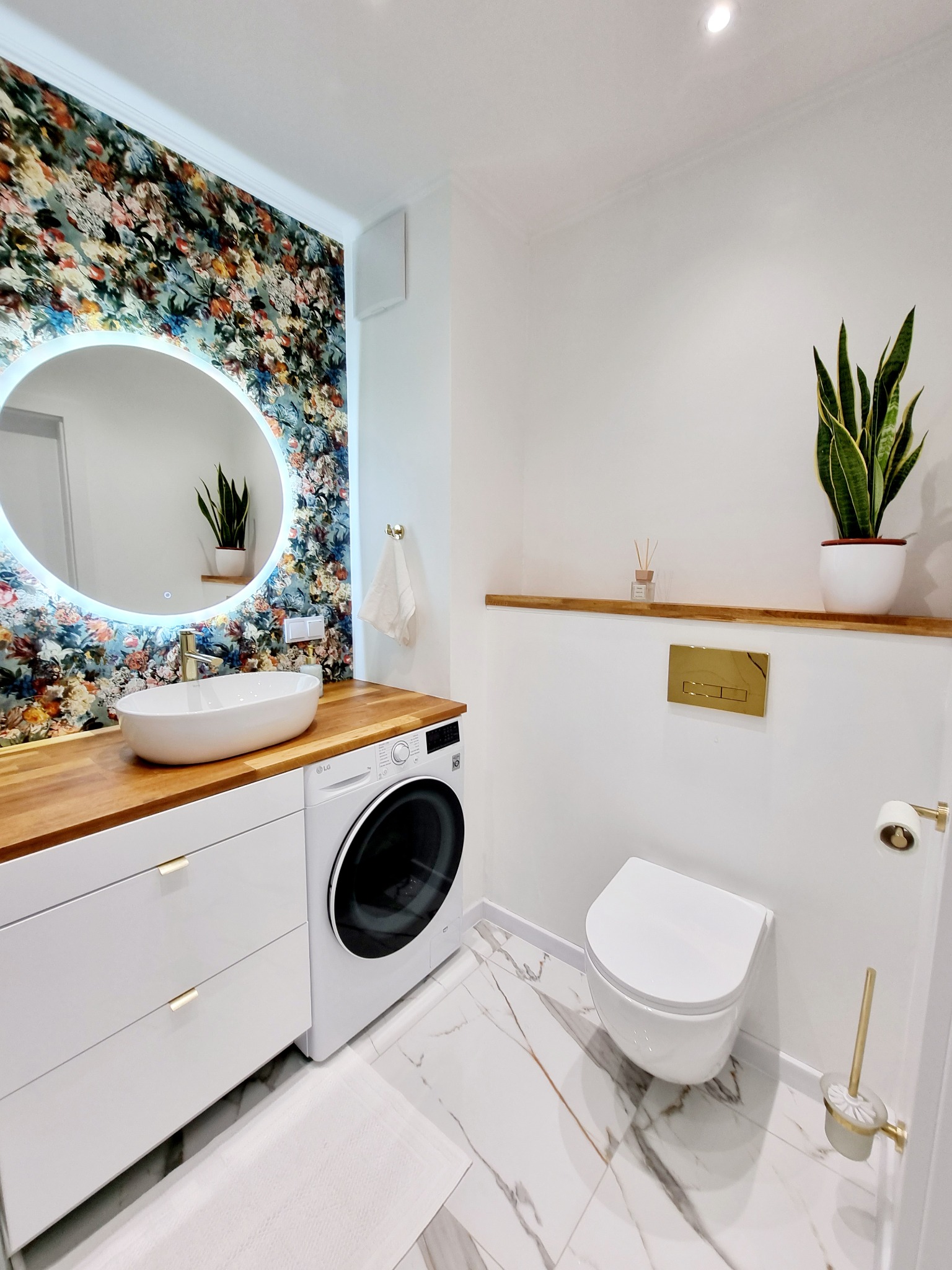 Nowoczesna łazienka z okrągłym lustrem LED, drewnianym blatem, białą umywalką i pralką. Marmurowe płytki podłogowe i ściana z motywem kwiatowym.