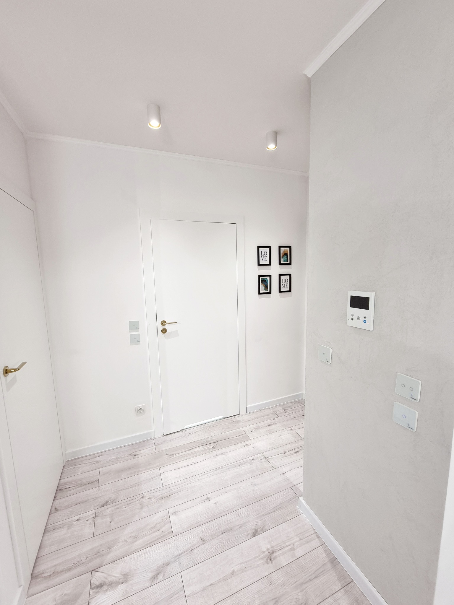 Jasne wnętrze korytarza z panelami podłogowymi imitującymi drewno, białymi ścianami z dekoracyjnym tynkiem, białymi drzwiami z złotymi klamkami i minimalistycznymi obrazkami.