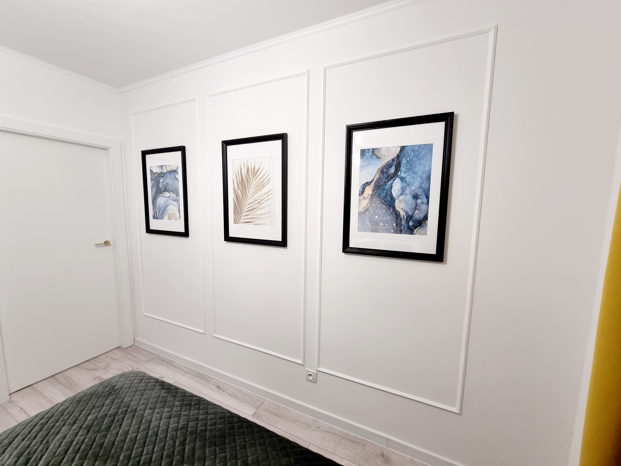 Białe ściany z dekoracyjnymi listwami i trzema obrazami w czarnych ramkach. Widoczny fragment szarego pufa i drzwi. Minimalistyczny design wnętrza z akcentami kolorystycznymi.