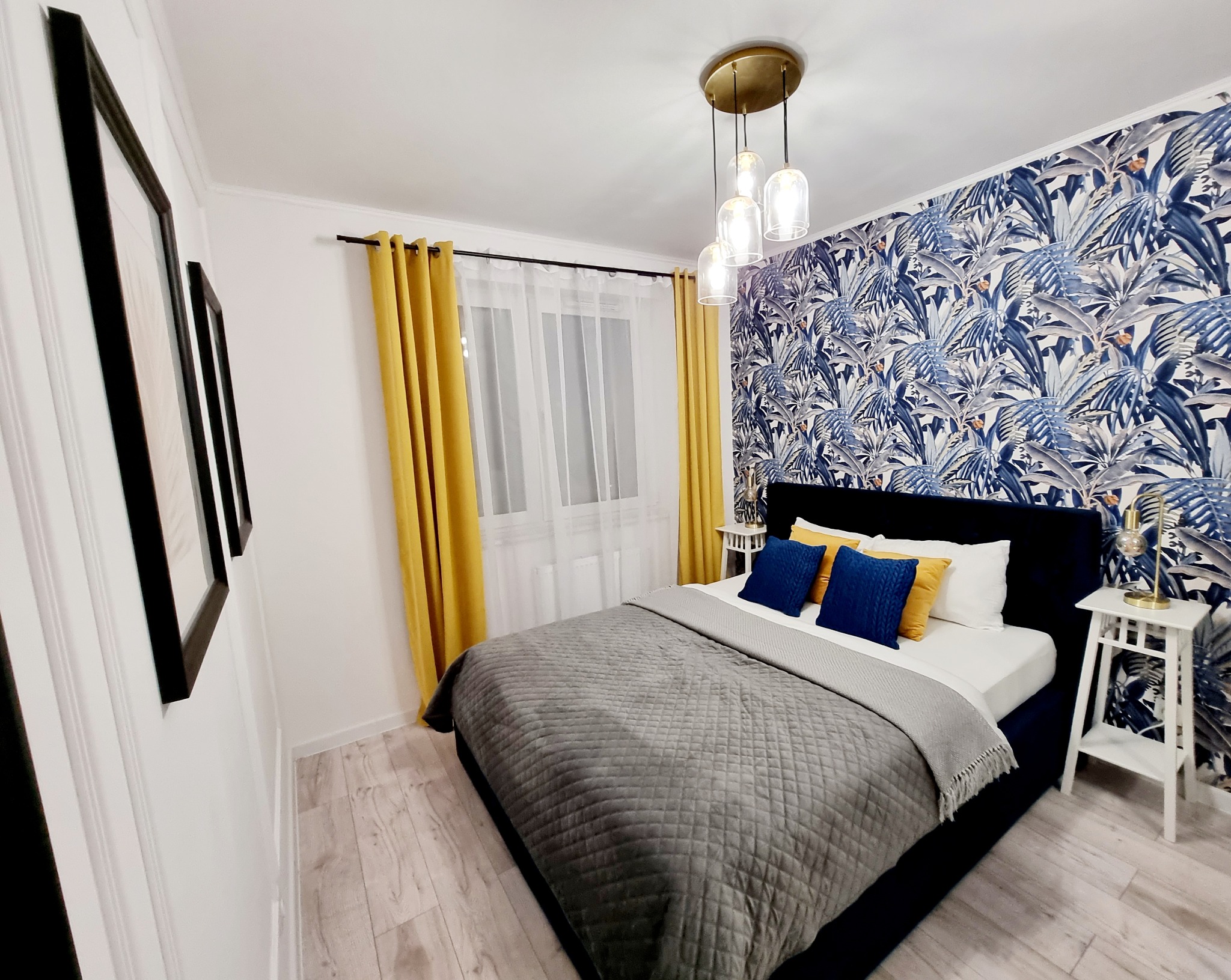 Nowoczesna sypialnia z tapetą w tropikalne liście, żółtymi zasłonami i szarą narzutą na łóżku. Widoczne drewniane panele podłogowe i lampa wisząca.