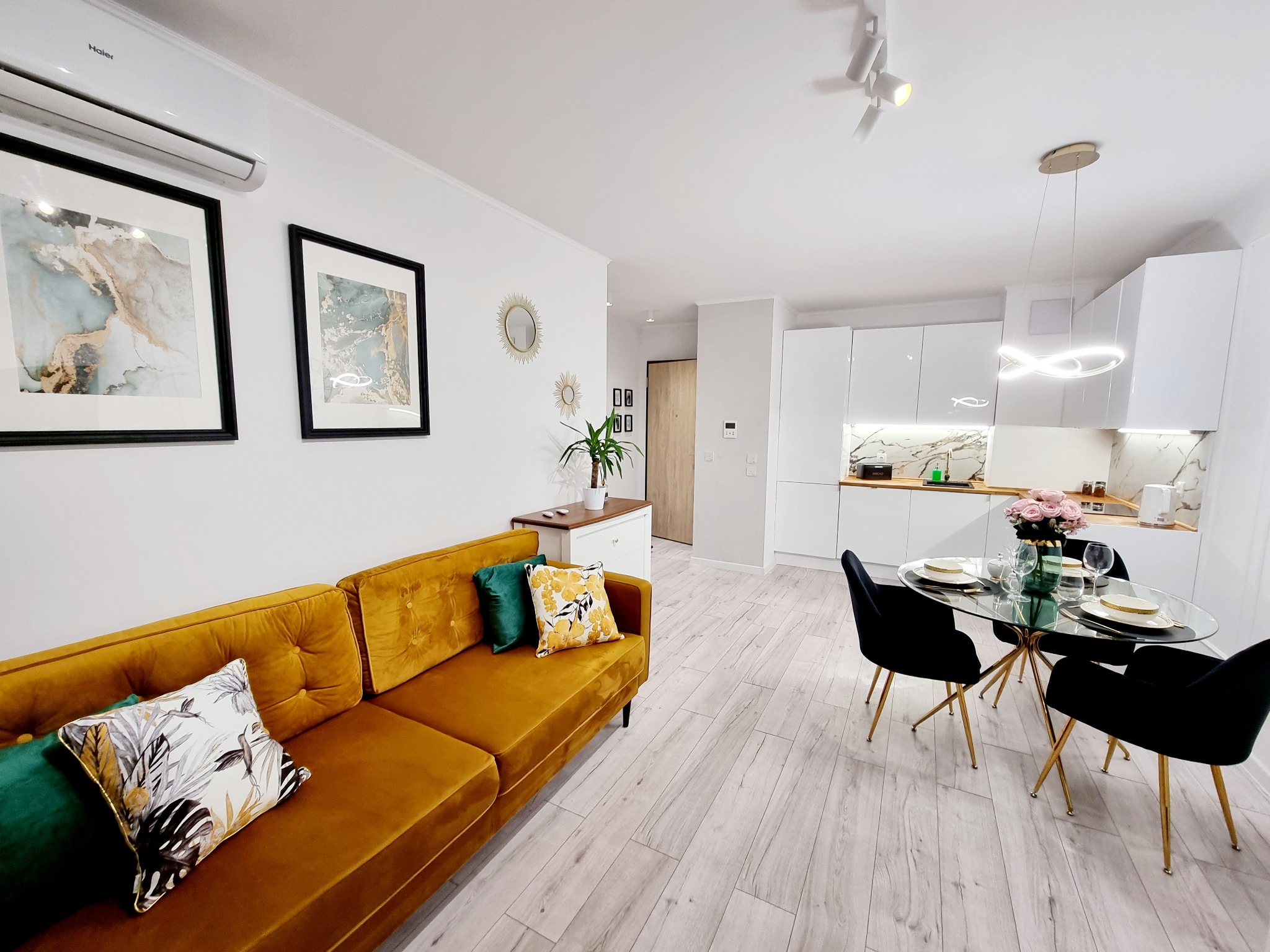 Jasne wnętrze po remoncie: złota sofa z poduszkami, stół z kwiatami, biała kuchnia z marmurowym blatem. Nowoczesne oświetlenie i drewnopodobna podłoga.