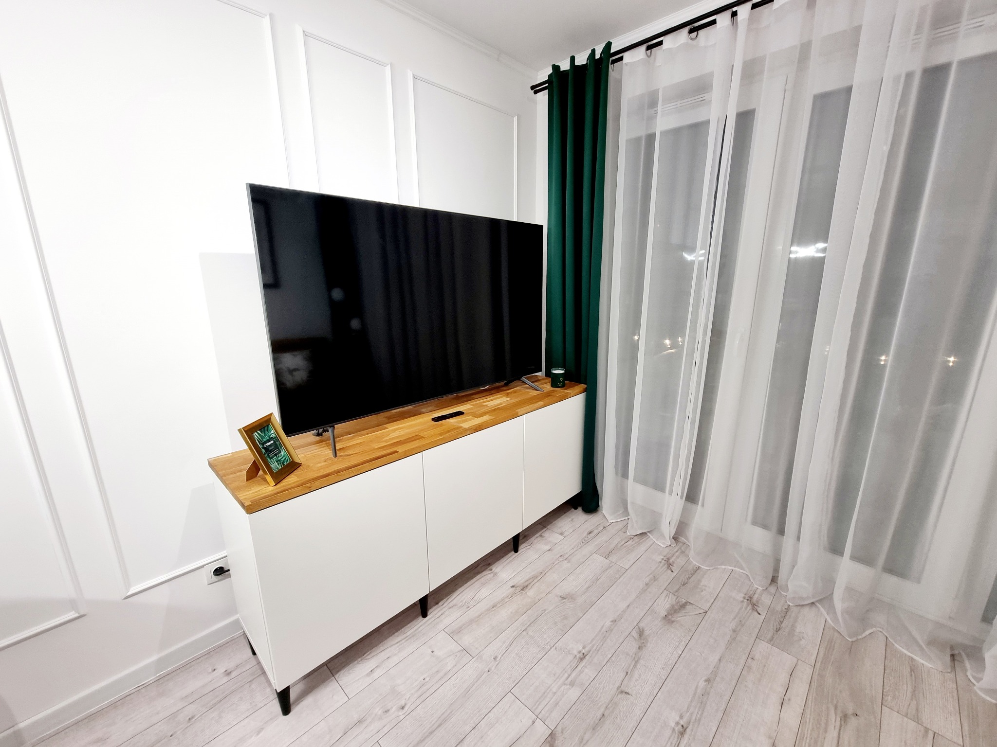 Nowoczesny salon z białą komodą z drewnianym blatem pod telewizorem, zielone zasłony i jasna podłoga. Ściana z dekoracyjnymi listwami.