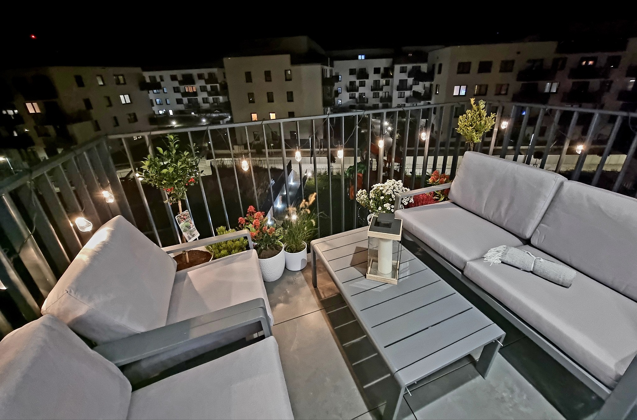 Nowoczesny balkon nocą: szare meble ogrodowe, rośliny w donicach, dekoracyjne lampki. Widok na oświetlone budynki w tle. Przytulna aranżacja balkonu.