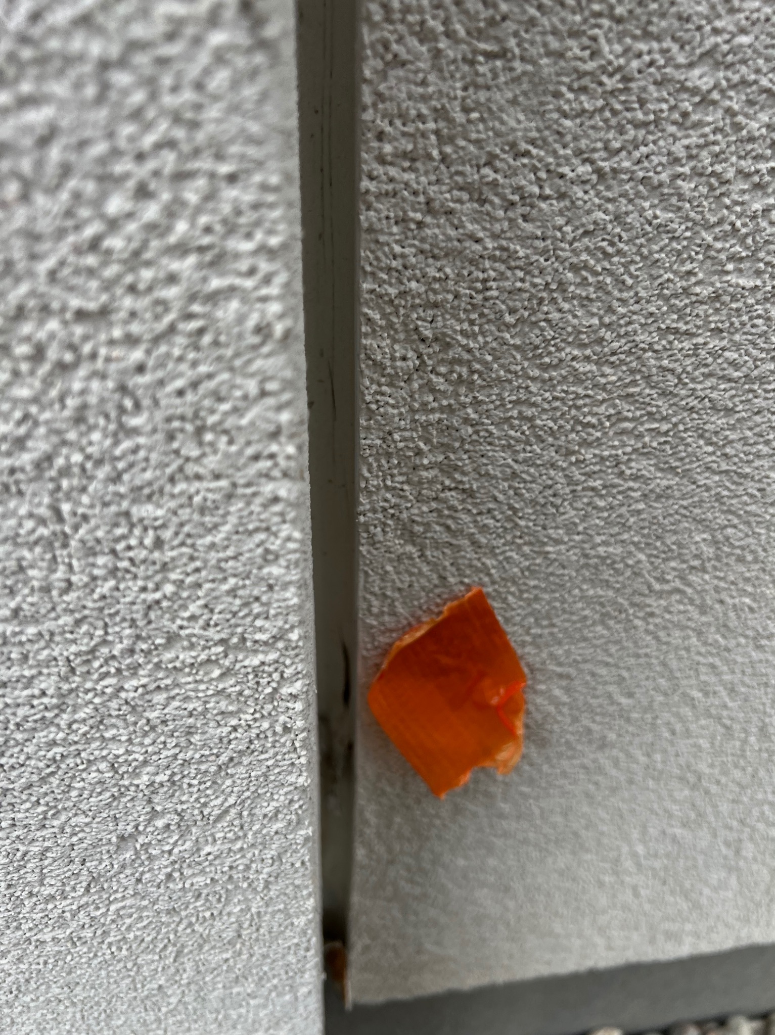 Fragment ściany z tynkiem barankiem, oddzielony pionową listwą. Na ścianie przyklejony pomarańczowy fragment taśmy malarskiej. Detal z inspekcji budowlanej.