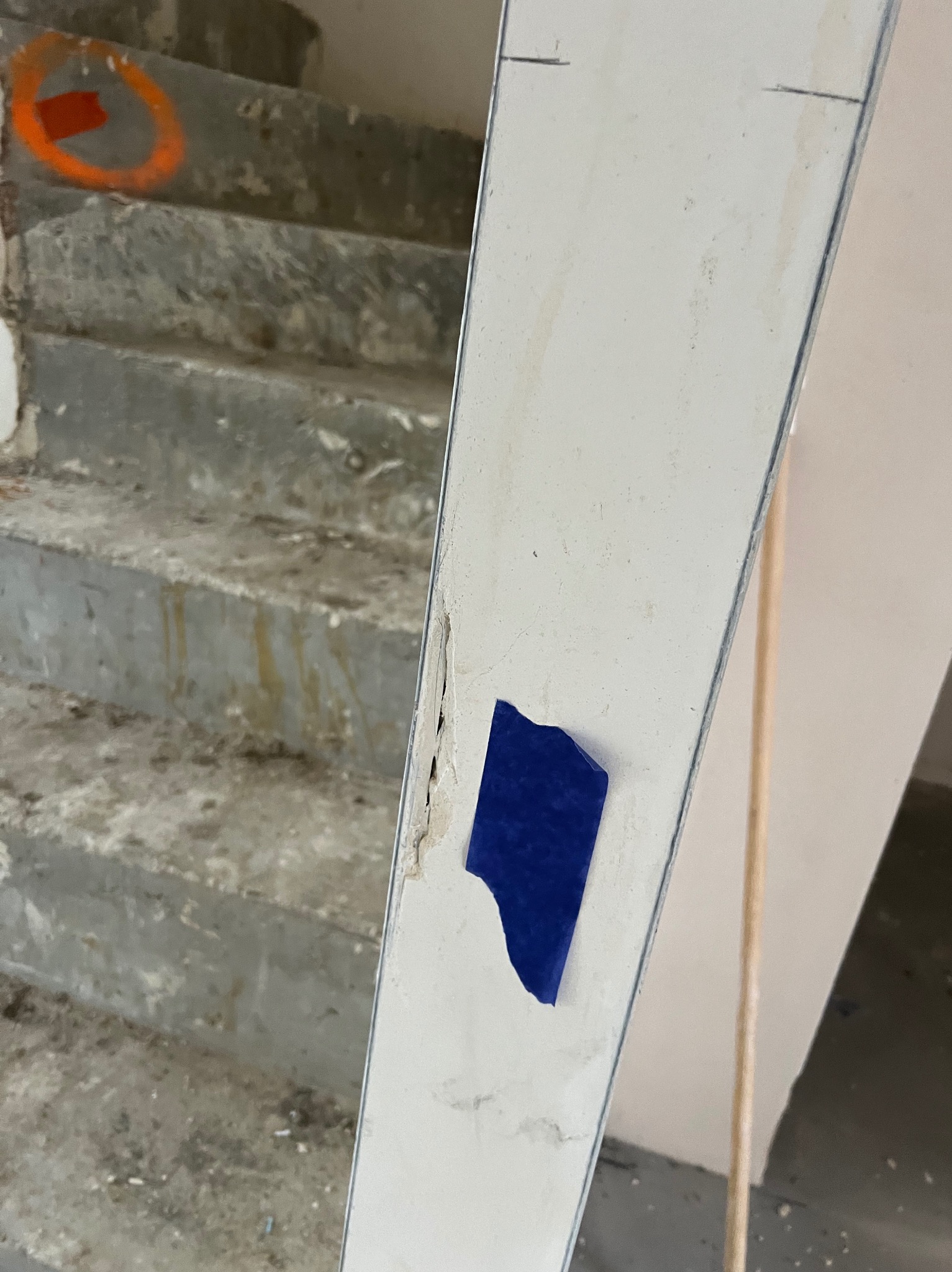 Niedoróbki wykończeniowe: uszkodzona ościeżnica z niebieską taśmą, widoczne schody z pomarańczowymi oznaczeniami. Detal z odbioru technicznego mieszkania.