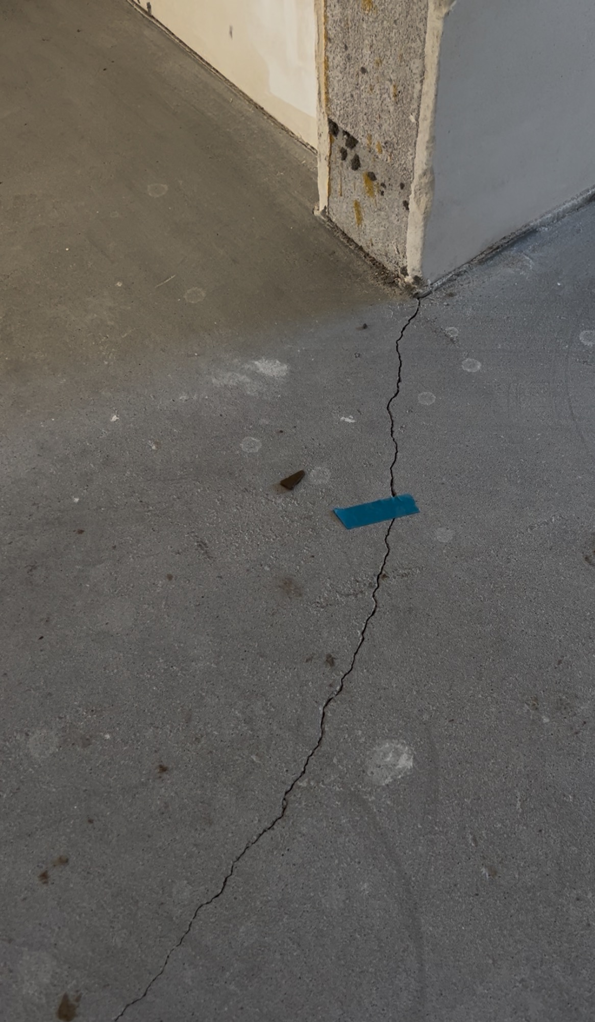 Szczelina w szarej, betonowej posadzce przy ścianie, oznaczona niebieską taśmą. Widoczne ślady po wierceniu i zabrudzenia. Ujęcie z bliska, podkreślające defekt.