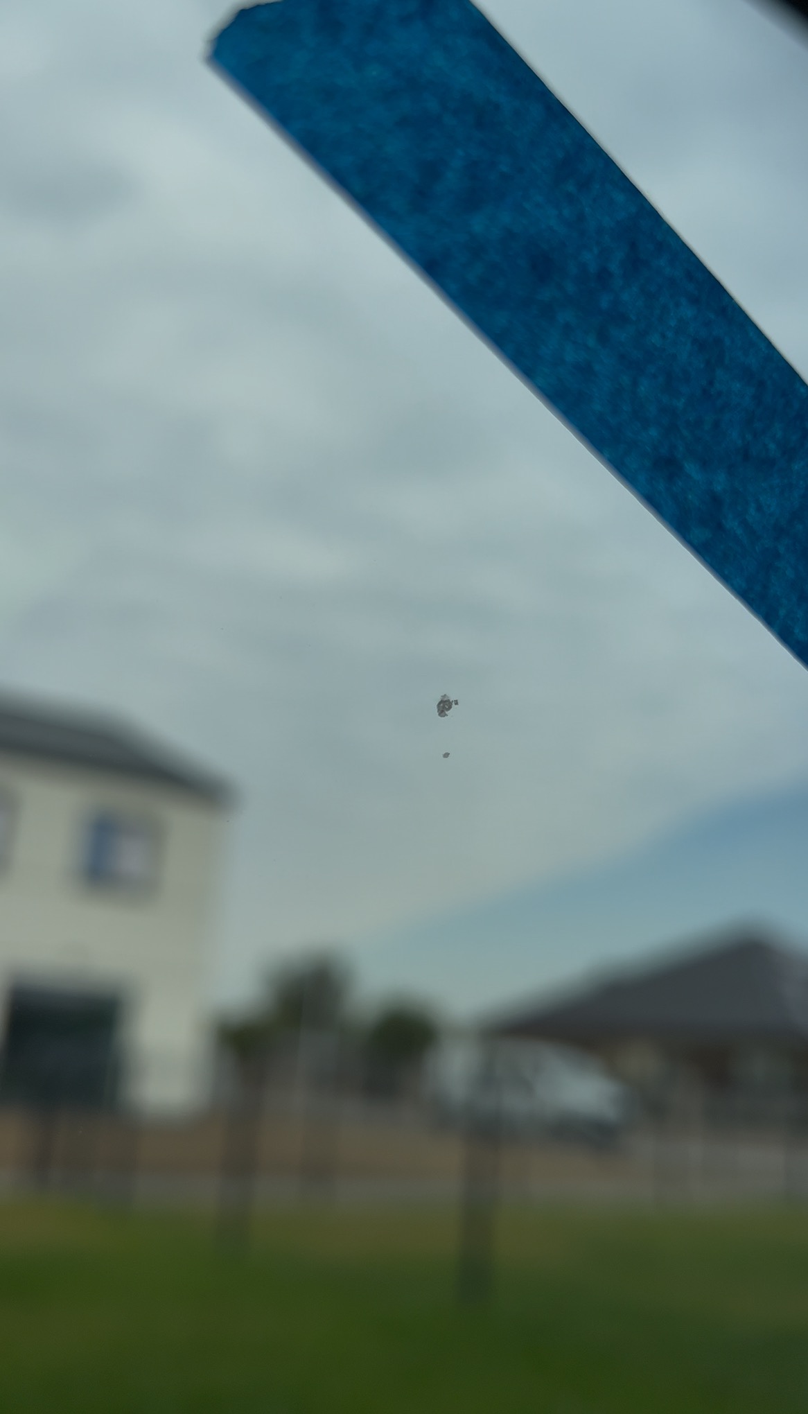 Drobne uszkodzenie szyby okiennej, zaznaczone niebieską taśmą, na tle rozmytego krajobrazu z domami. Inspekcja jakości wykonania okien.