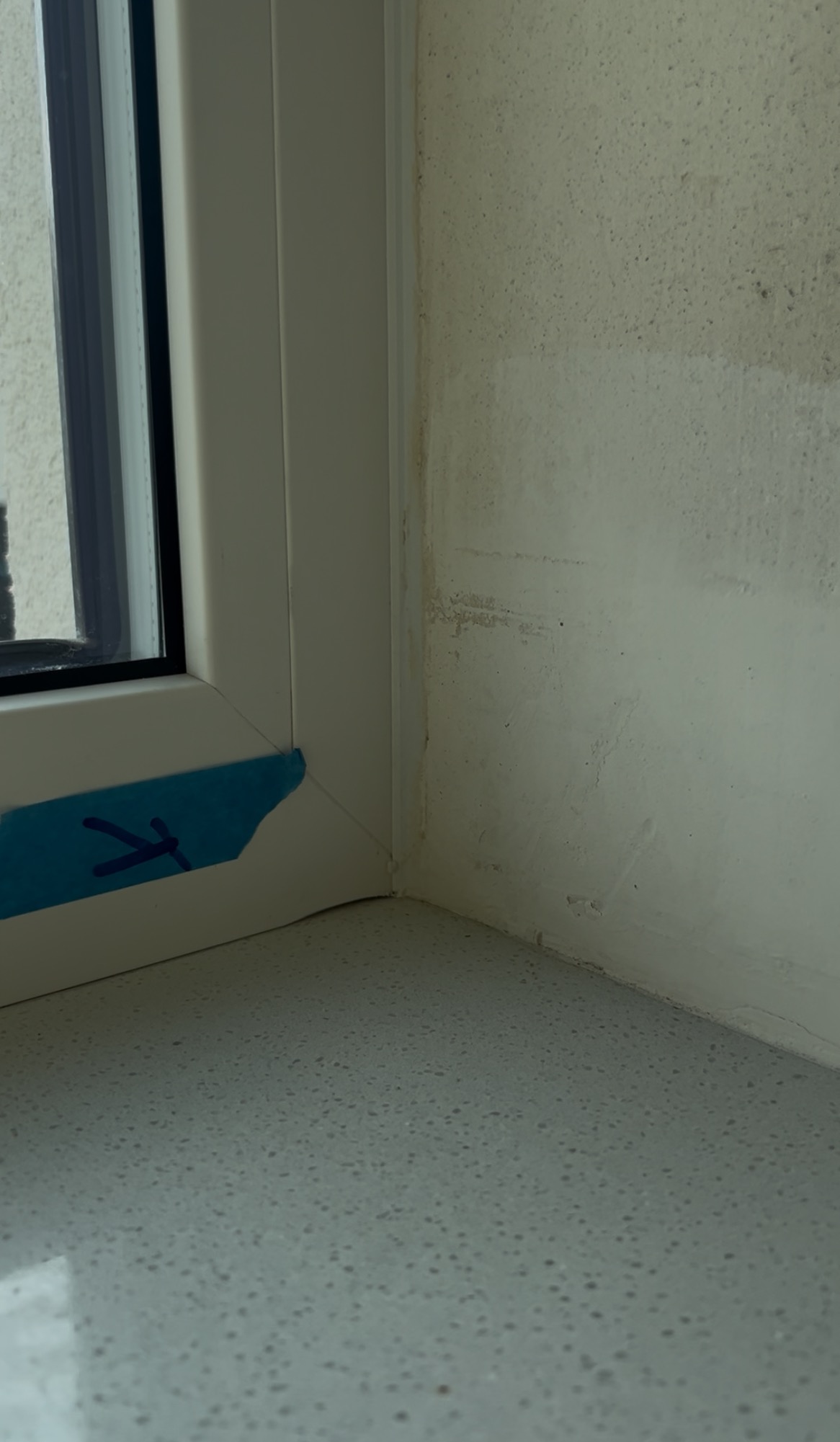 Narożnik okna z taśmą i strzałką wskazującą na zawilgocenia i pleśń na ścianie. Widoczne niedociągnięcia podczas odbioru mieszkania.