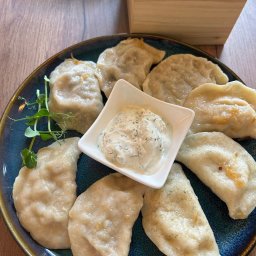 Yuliia Ivchenko - Ręcznie lepione pierogi z sosem na niebieskim talerzu, Oświęcim. Dekoracja z zielonych pędów. Drewniany stół i pojemnik na przyprawy w tle. Bliski kadr.