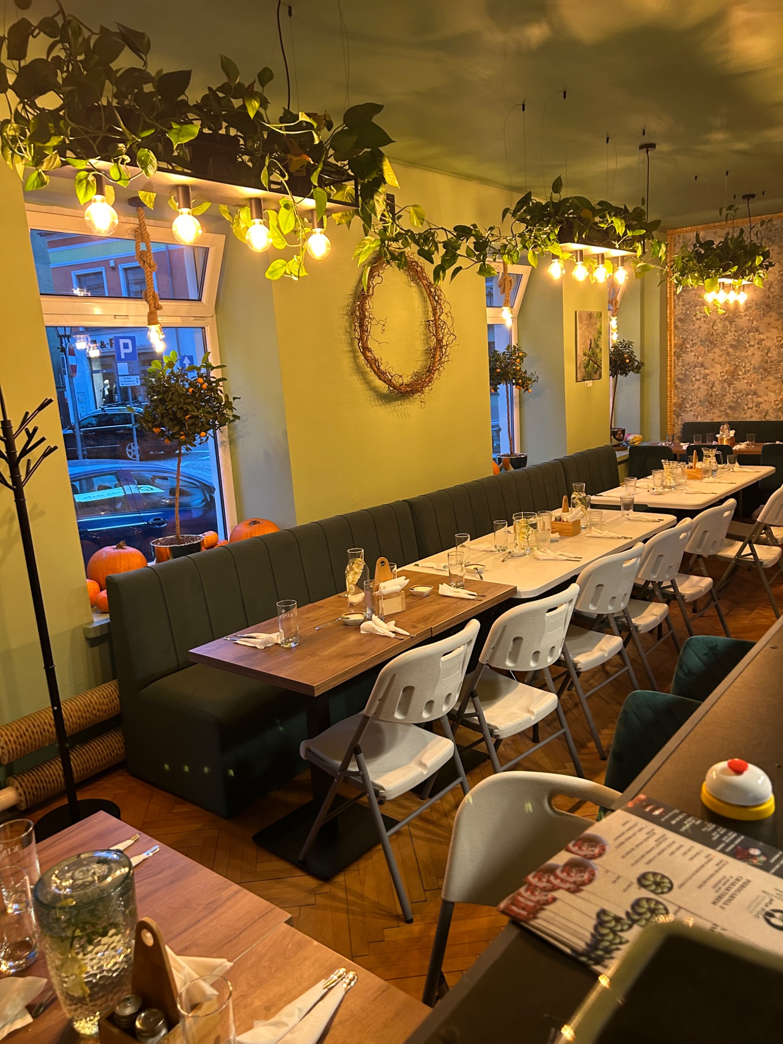 Ustawione stoły w restauracji z zielonymi ścianami i roślinnymi dekoracjami. Na oknach dynie, a na stołach nakrycia i szkło. Ciepłe oświetlenie tworzy przytulną atmosferę.