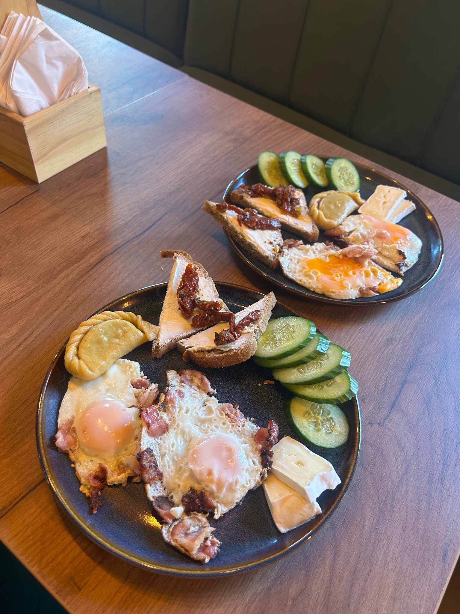 Dwa talerze z różnorodnymi przekąskami: jajka sadzone z boczkiem, kanapki z suszonymi pomidorami, ogórki i ser camembert na drewnianym stole.