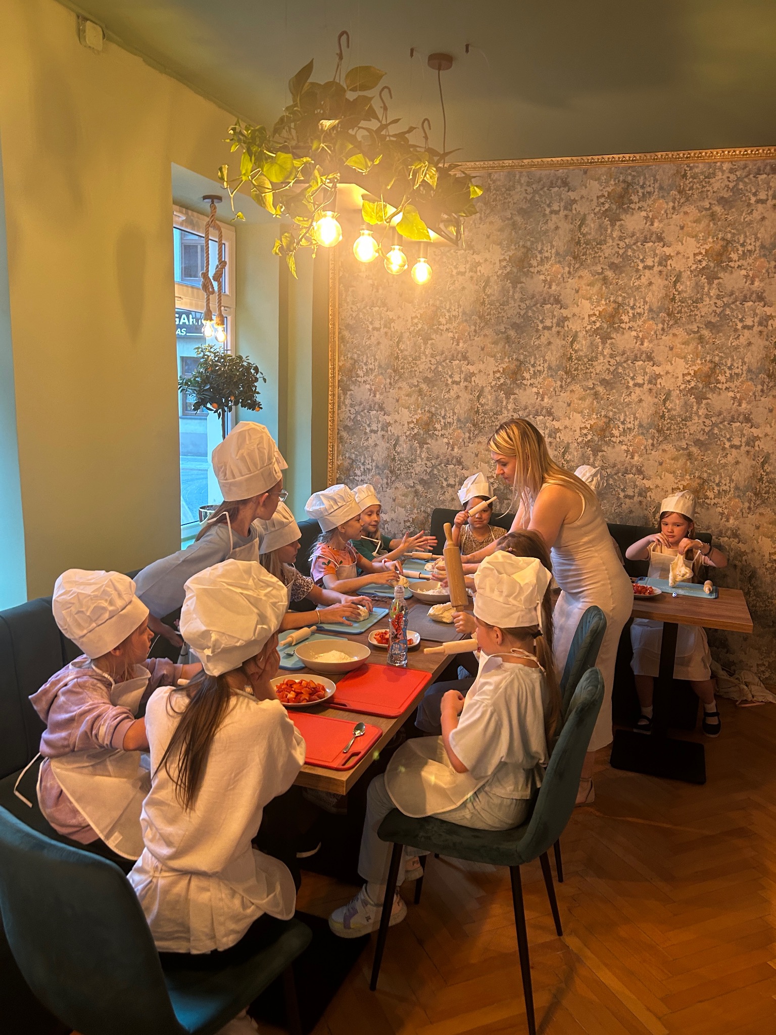 Warsztaty kulinarne dla dzieci: grupa w czapkach kucharskich przygotowuje posiłek pod okiem instruktorki w jasnym wnętrzu z dekoracyjną ścianą i roślinami.