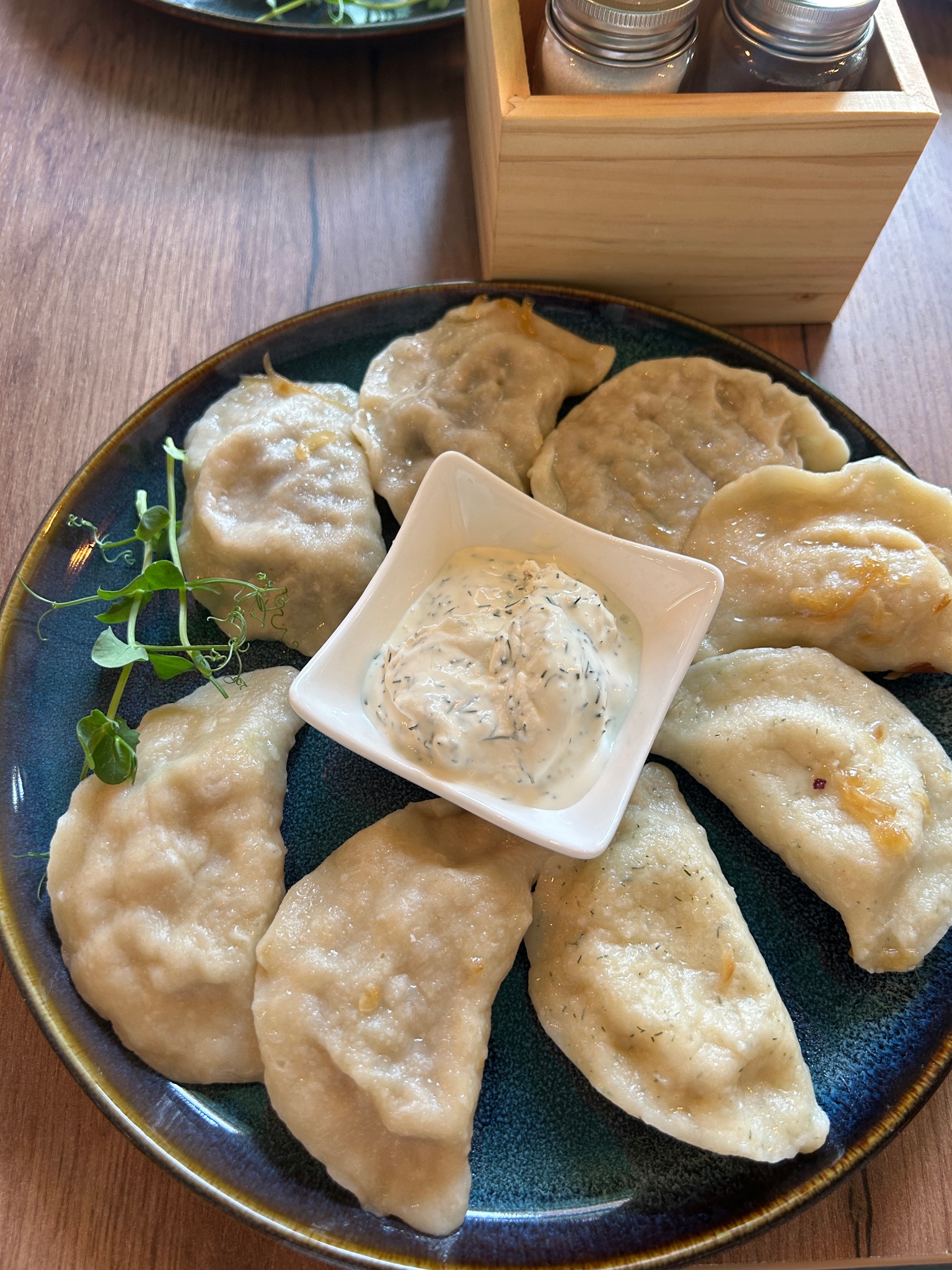 Ręcznie lepione pierogi z sosem na niebieskim talerzu, Oświęcim. Dekoracja z zielonych pędów. Drewniany stół i pojemnik na przyprawy w tle. Bliski kadr.