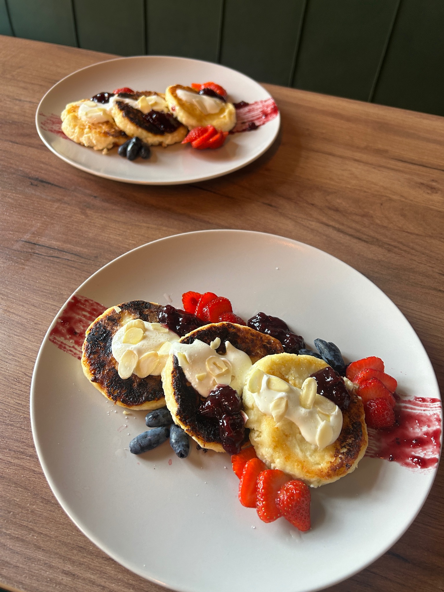 Dwa talerze z sernikami na słodko, udekorowane bitą śmietaną, dżemem i świeżymi truskawkami oraz jagodami na drewnianym stole. Widok z góry, ciepłe światło.