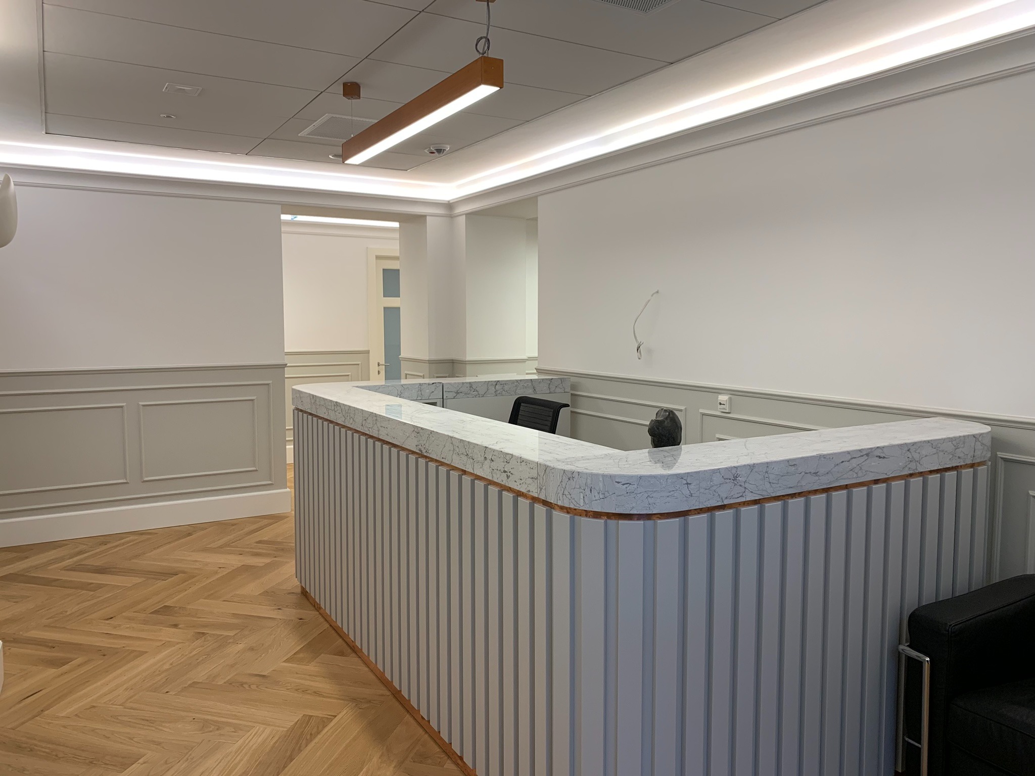 Wykończone wnętrze biura: recepcja z blatem imitującym marmur, panele ścienne w odcieniach szarości, parkiet w jodełkę i oświetlenie LED. Nowoczesny design.