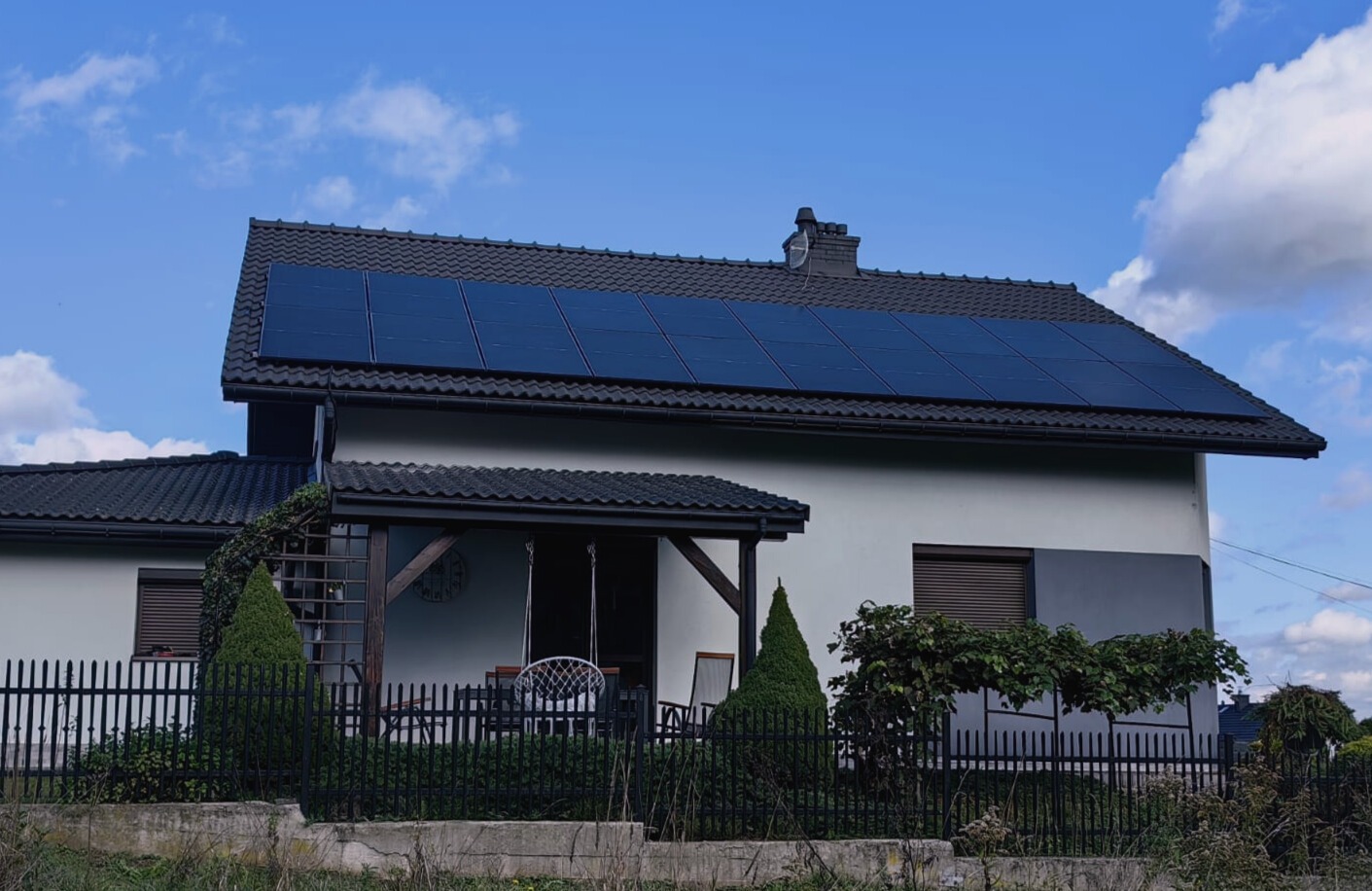 Dom jednorodzinny z panelami słonecznymi na dachu, widok zza czarnego ogrodzenia. Jasna elewacja, ciemna dachówka i stolarka okienna. Błękitne niebo z chmurami w tle.