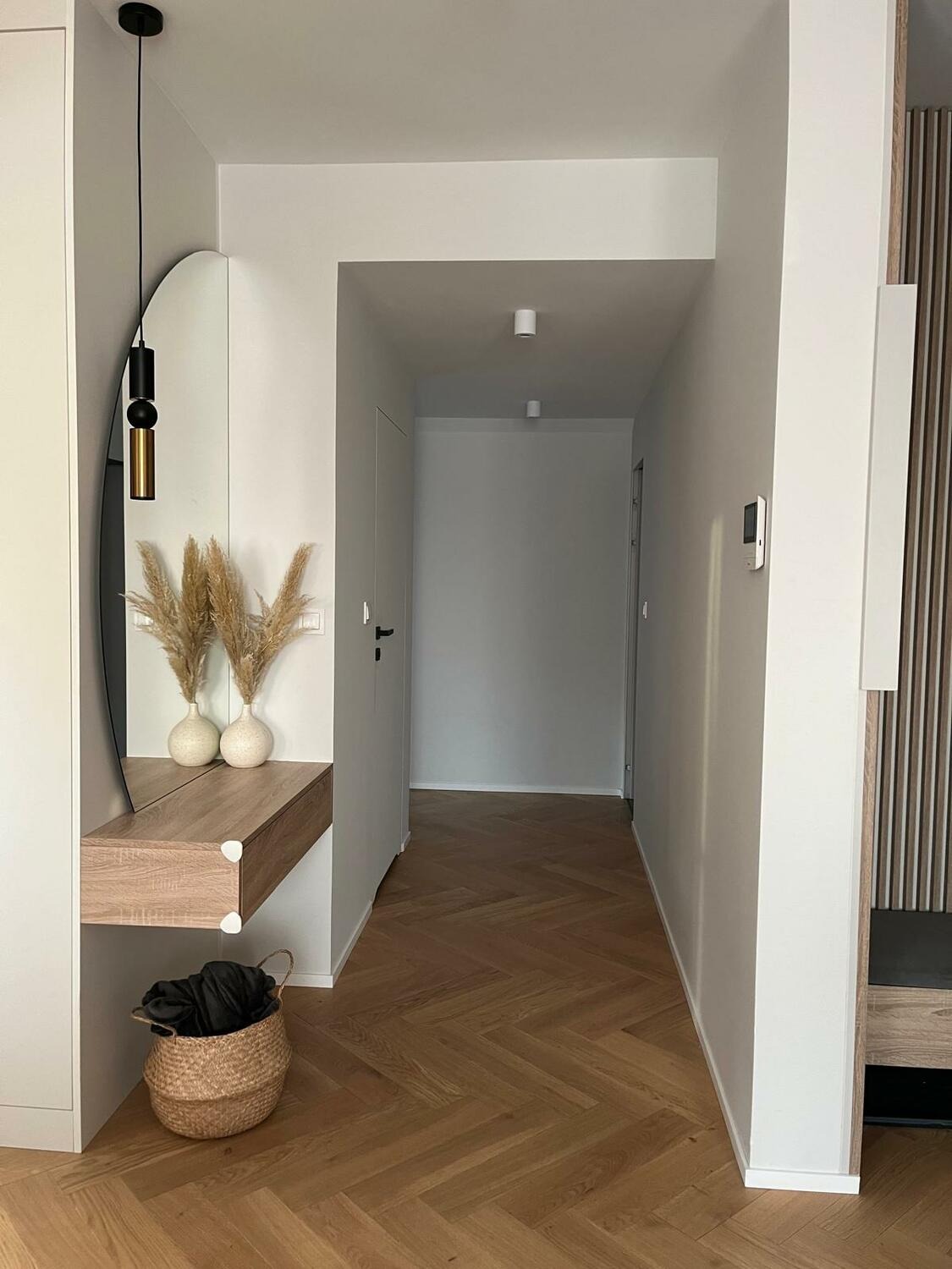 Nowoczesny przedpokój z lustrem, drewnianą konsolą i parkietem w jodełkę. Minimalistyczne oświetlenie i dekoracyjne trawy pampasowe dodają wnętrzu elegancji.