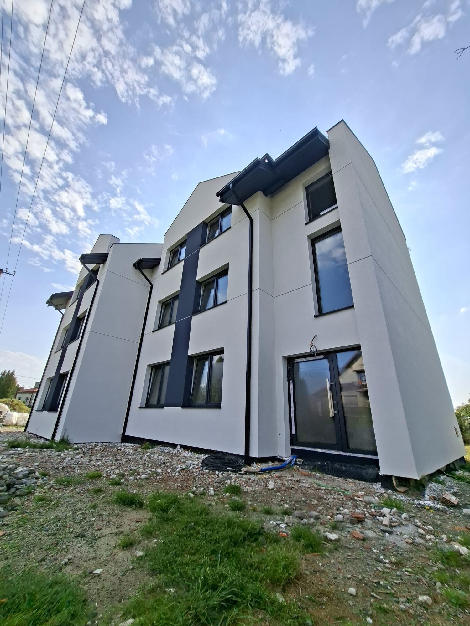 Nowoczesny budynek po remoncie elewacji z grafitowymi akcentami, widok z dołu na tle błękitnego nieba z chmurami. Teren wokół budynku jeszcze niezagospodarowany.