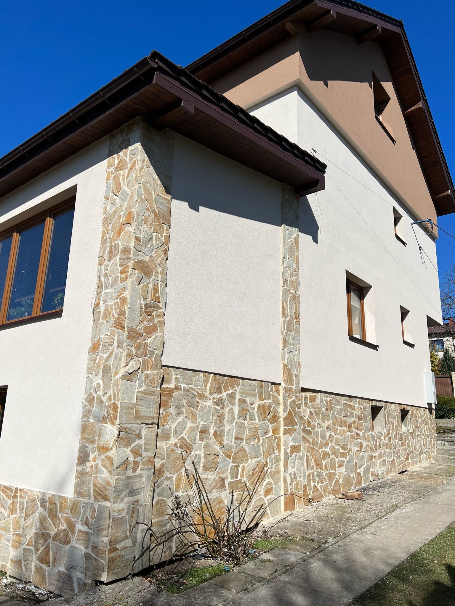 Wykończenie elewacji domu z elementami kamiennymi i jasnym tynkiem. Widoczne okna z brązowymi ramami i fragment dachu. Słoneczny dzień, błękitne niebo.