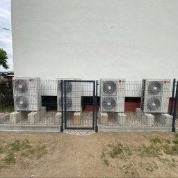 Krople Ciepła - Sześć jednostek zewnętrznych pomp ciepła LG Therma V na betonowych cokołach, zabezpieczonych czarnym ogrodzeniem panelowym z furtką, przy białej elewacji budynku.