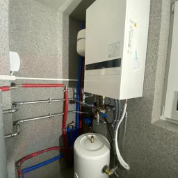 Krople Ciepła - Nowoczesna instalacja grzewcza z białą pompą ciepła na ścianie i zasobnikiem na podłodze, z widocznymi rurami w czerwonej i niebieskiej izolacji, w pomieszczeniu gospodarczym z szarymi...
