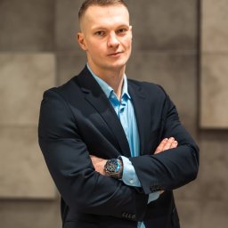 A.S. Consulting Sp. Z o.o. - Mężczyzna w garniturze, z zegarkiem na ręku, stoi na tle ściany z szarych paneli. Uśmiechnięty, pewny siebie, z założonymi rękami. Profesjonalny wizerunek.