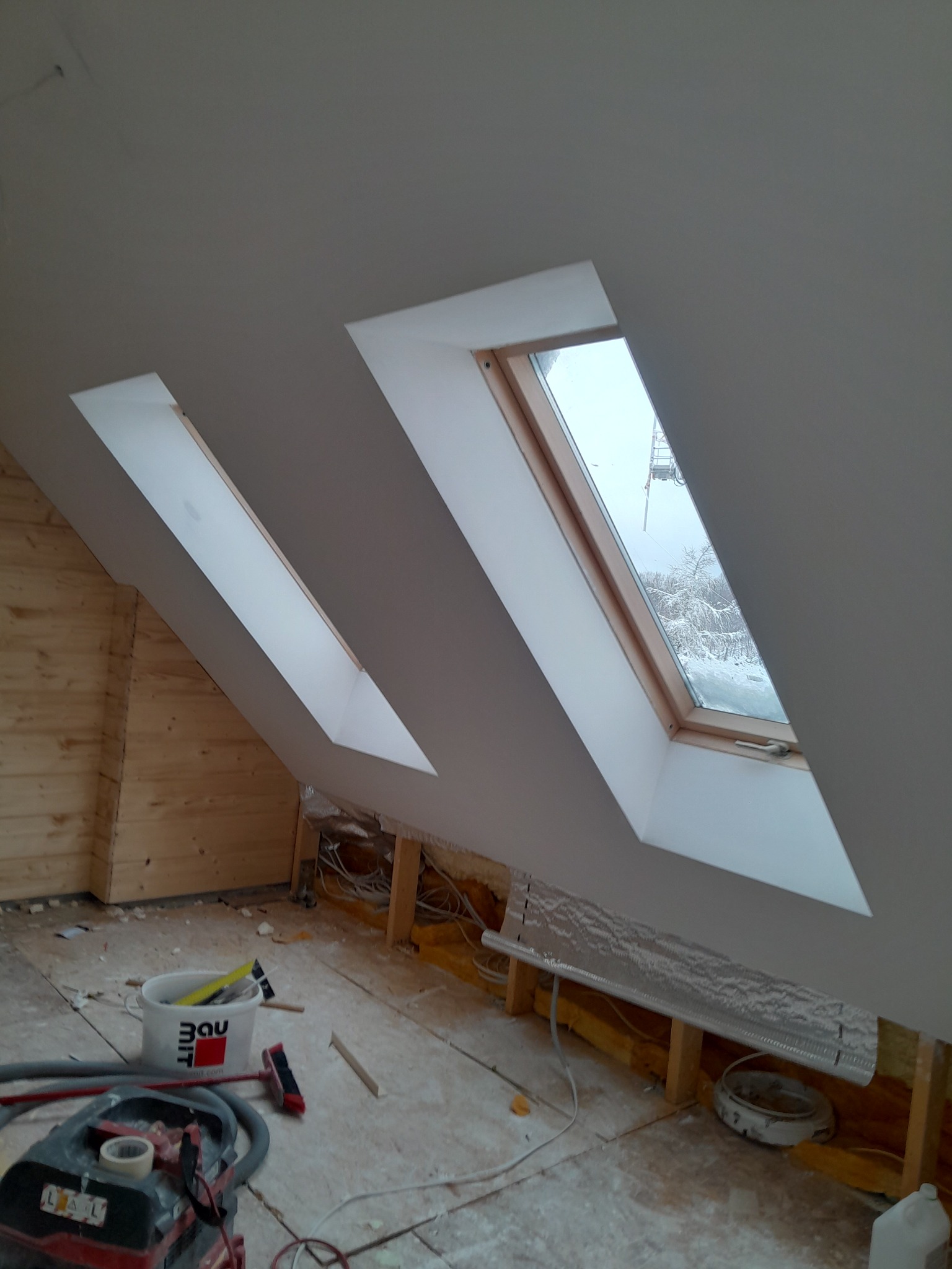 Poddasze po montażu okien dachowych i wykończeniu płytami gipsowo-kartonowymi. Widoczne narzędzia i materiały budowlane. Śnieżny krajobraz za oknem.