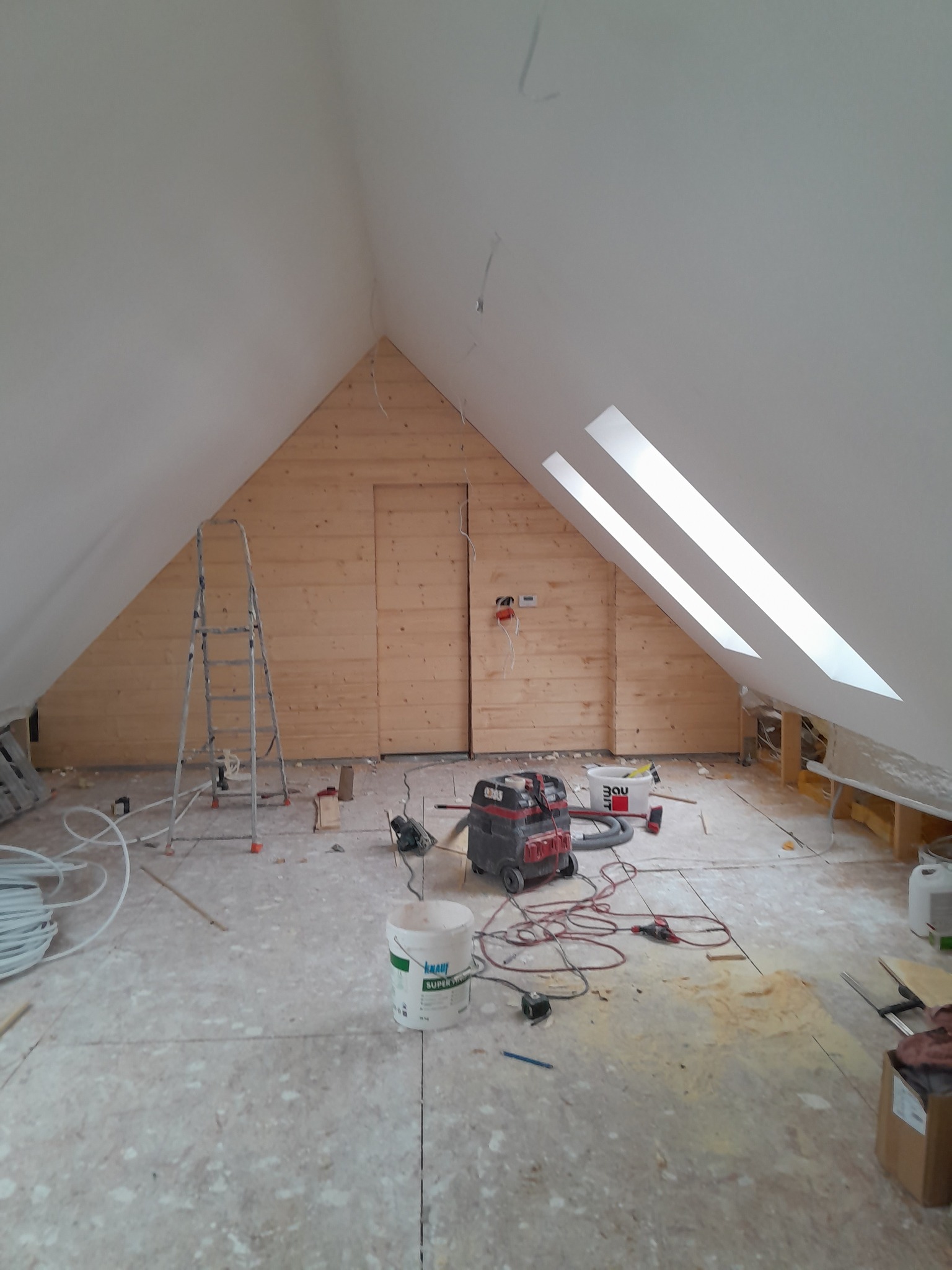 Poddasze w trakcie prac wykończeniowych: drewniana boazeria na ścianie szczytowej, okna dachowe, drabina, narzędzia i materiały budowlane na podłodze. Widok z dołu.