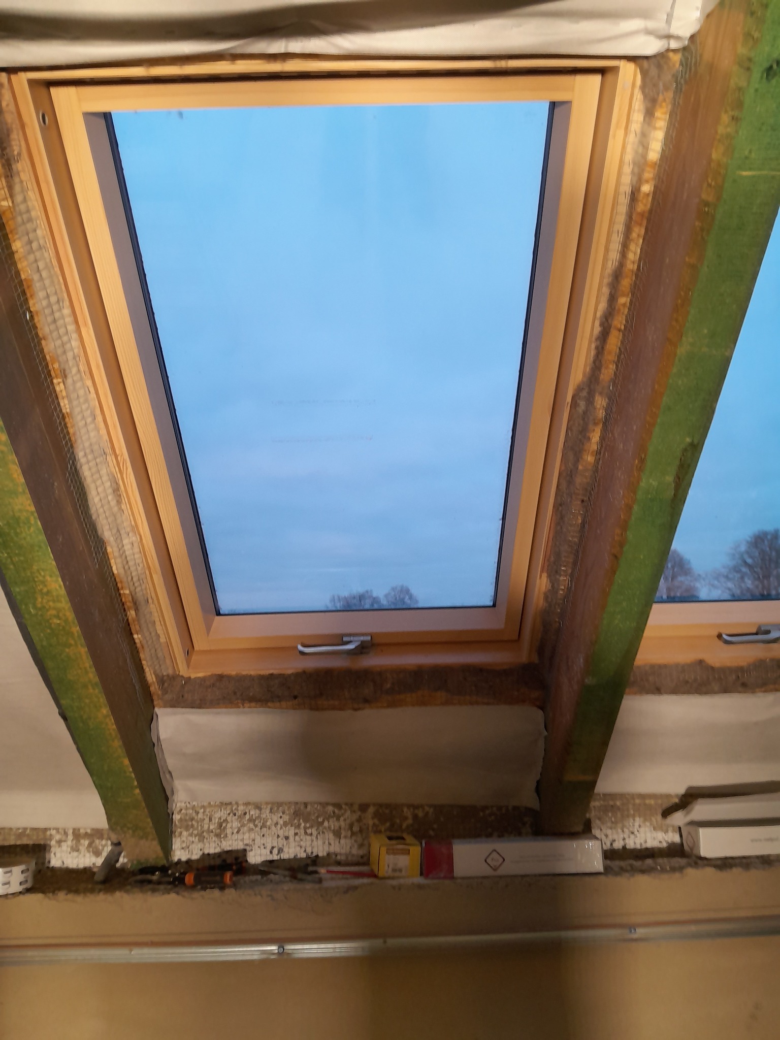 Montaż okna dachowego z drewnianą ramą, widoczne elementy konstrukcyjne dachu i izolacja. Ujęcie z dołu, w tle błękitne niebo. Widoczne narzędzia i materiały budowlane.