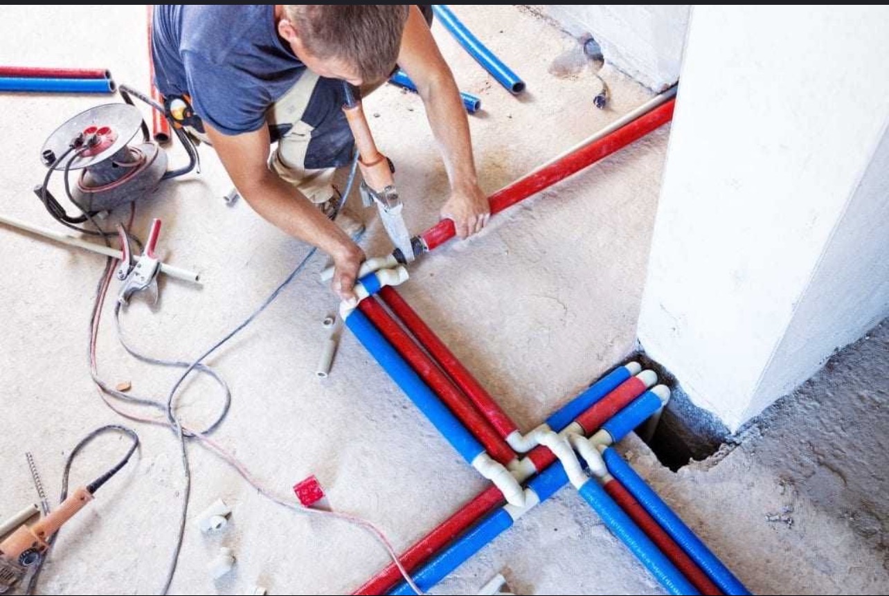 Instalacja rur PEX: hydraulik łączy czerwone i niebieskie rury złączkami. Widoczne narzędzia i przewody. Praca w toku, surowe wykończenie podłoża.