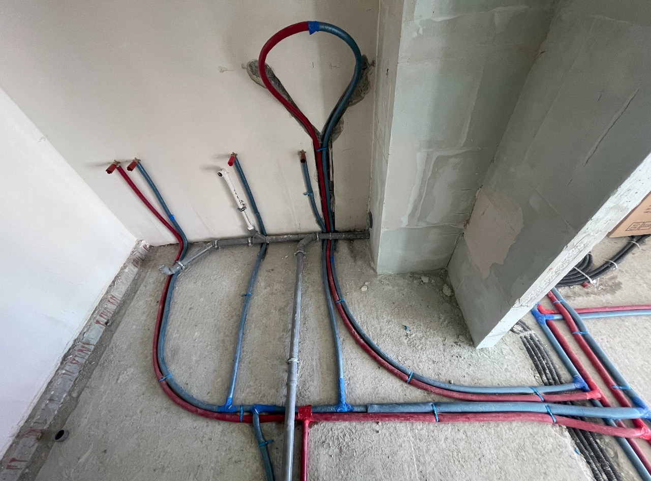 Instalacja wodna i grzewcza w surowym stanie: rury PEX czerwone i niebieskie prowadzone po ścianach i podłodze, widoczne złącza i mocowania.