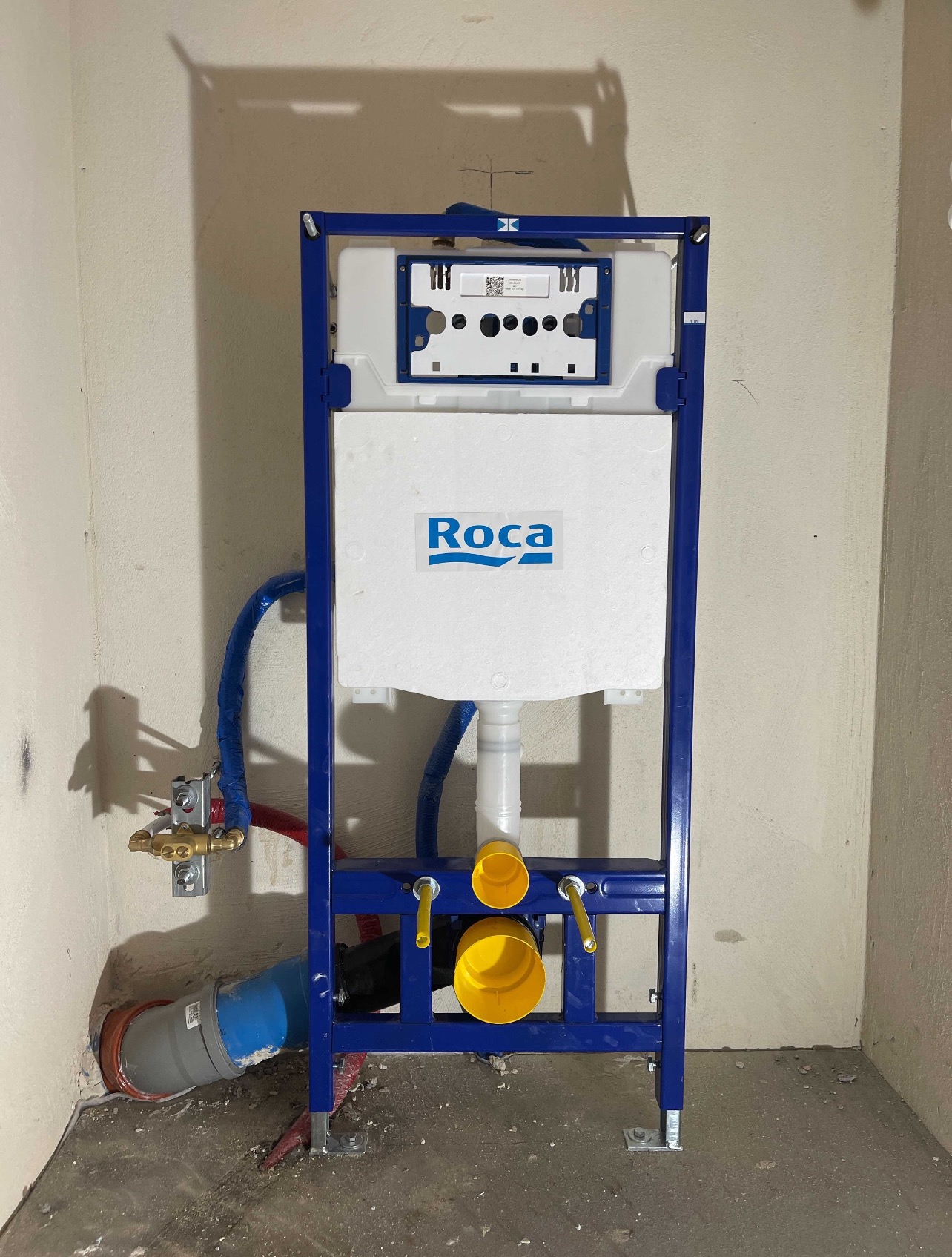 Stelaż podtynkowy WC marki Roca w trakcie montażu, widoczne przyłącza wodne i kanalizacyjne. Surowe ściany i podłoga wskazują na etap prac wykończeniowych.