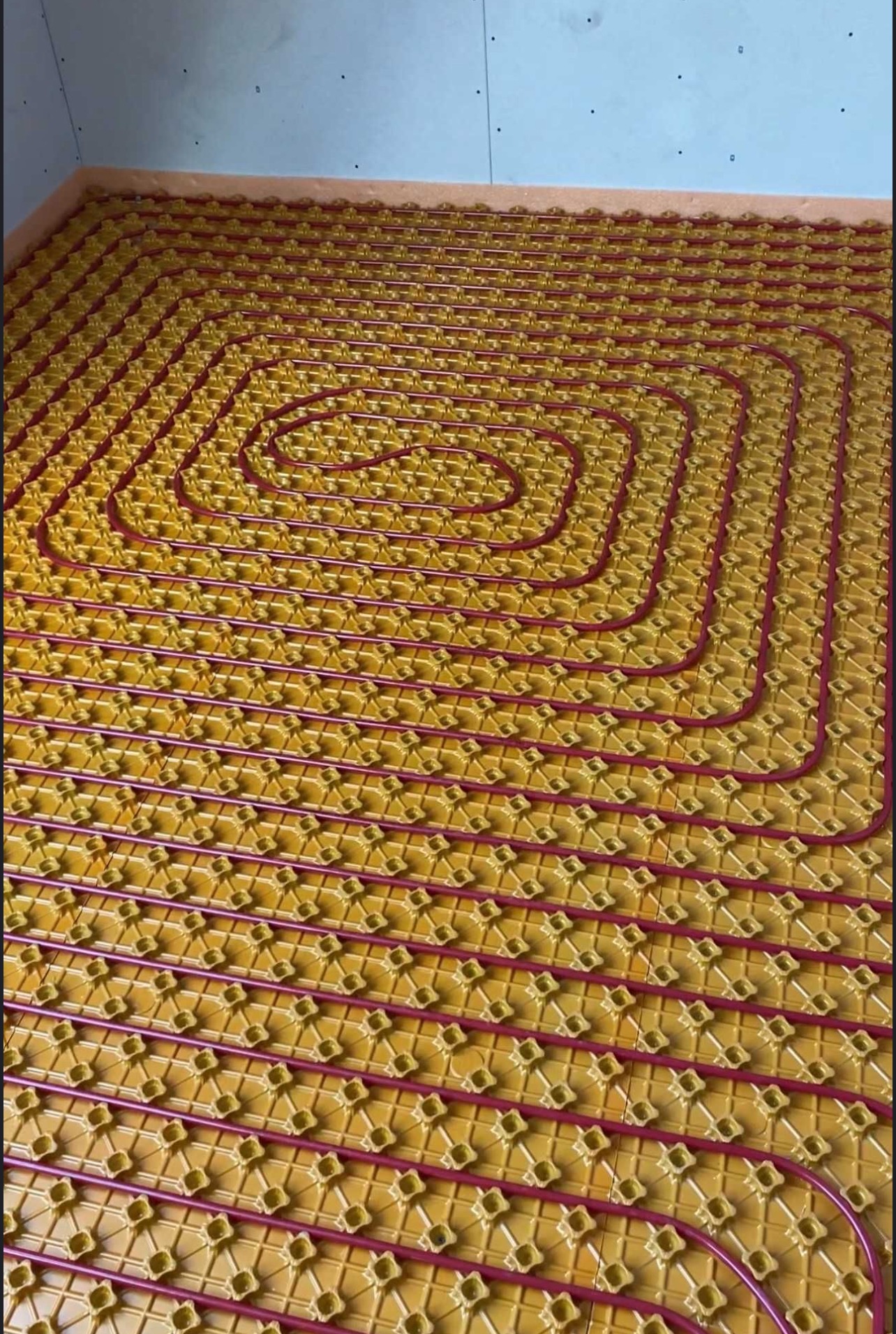 Żółte maty z czerwonymi rurkami ogrzewania podłogowego ułożone spiralnie na przygotowanej powierzchni. Widok z góry, bliski kadr.